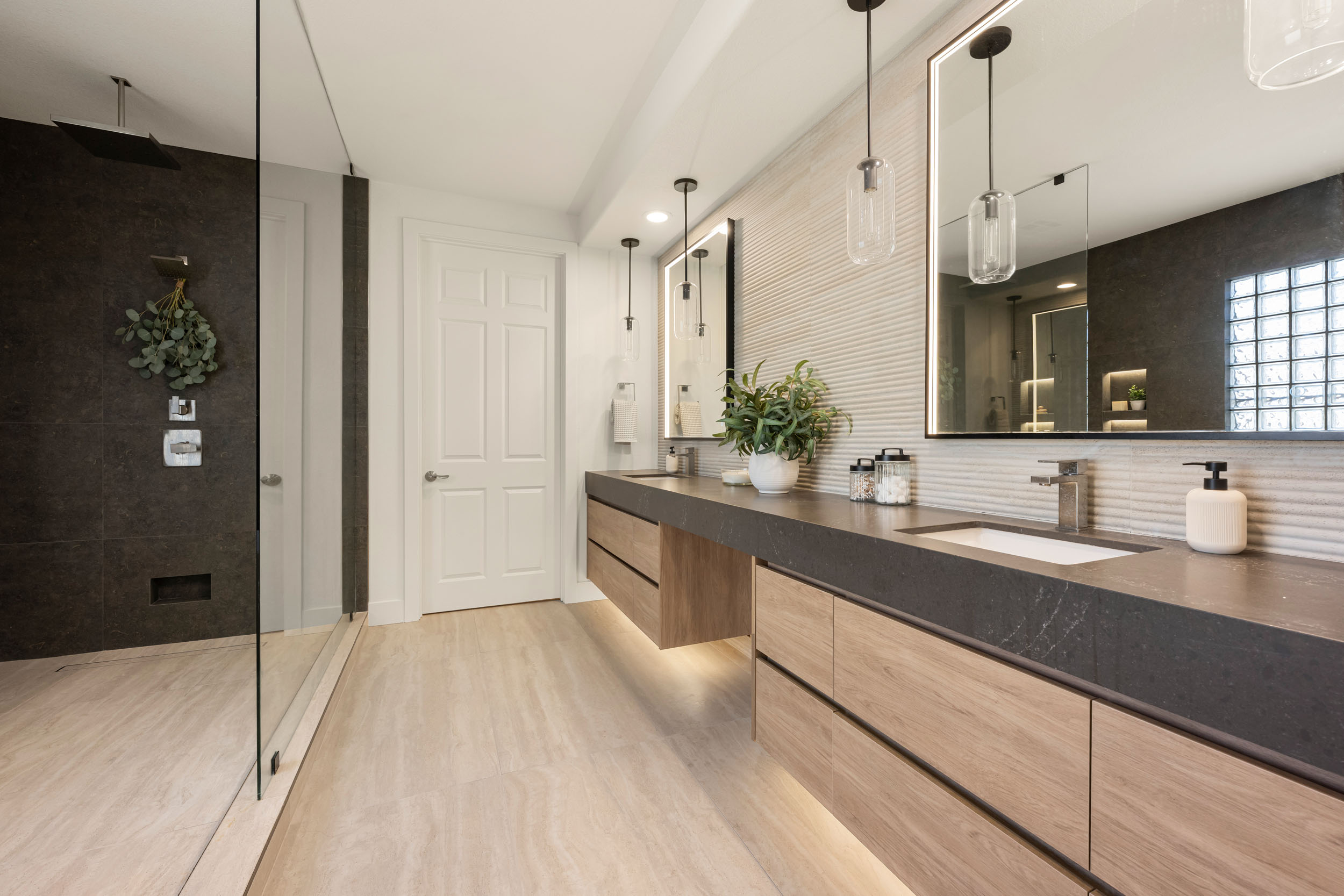 Primary bathroom wide with floating oak double vanity, honed dark-stone counter, glass cylinder pendants, and ribbed tile backsplash in Summerlin remodel by Kingdom & Co.