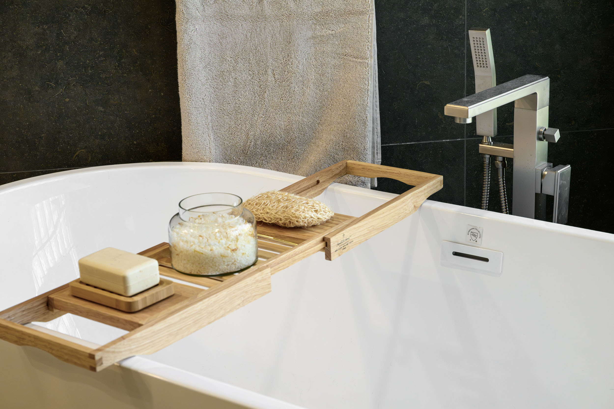 Freestanding white soaker tub with wood bath caddy and wall-mount brushed nickel tub filler against dark stone wall in Summerlin remodel by Kingdom & Co.