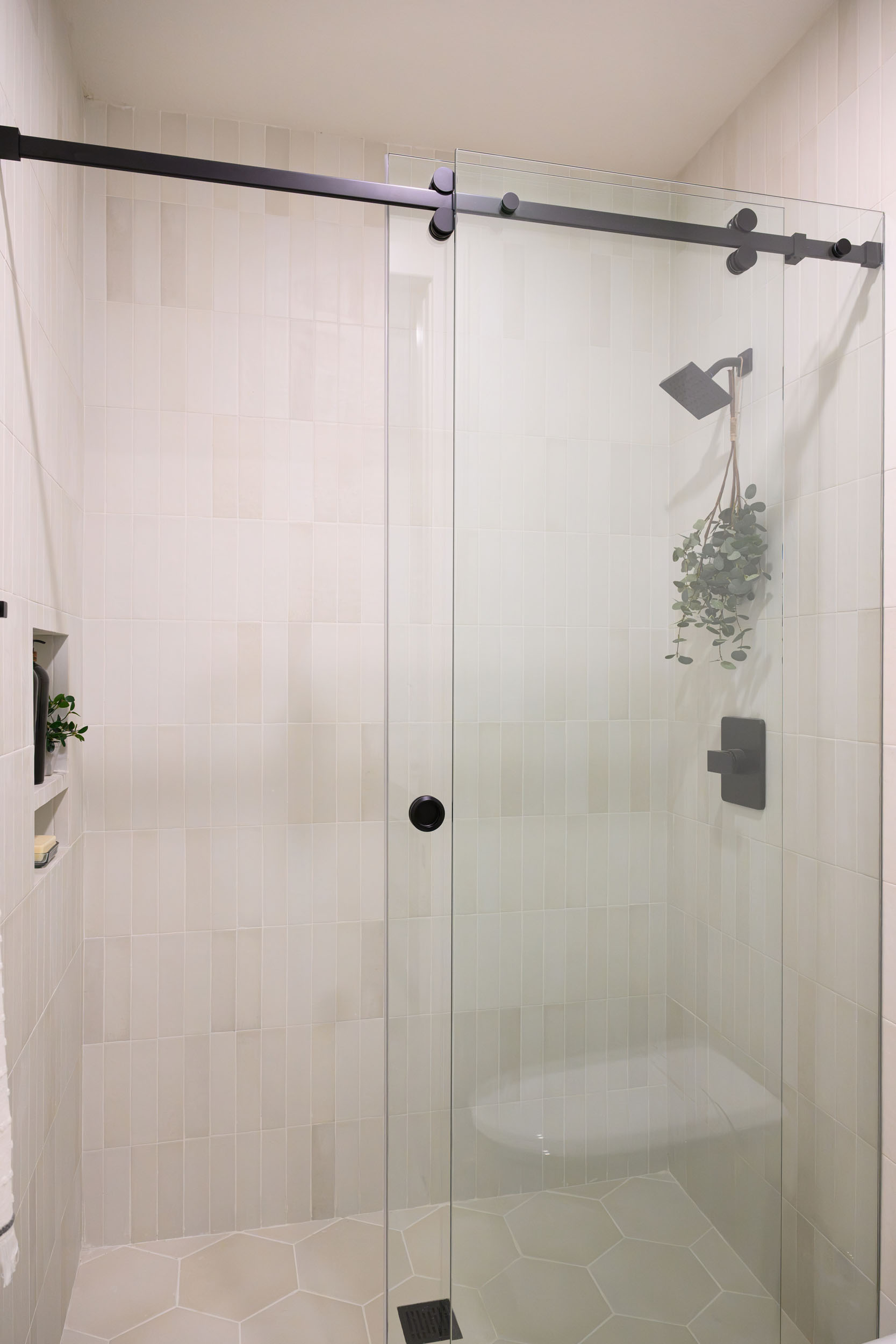 Secondary shower with black-framed sliding glass door on stacked vertical white tile walls and hex-tile floor in Summerlin remodel by Kingdom & Co.